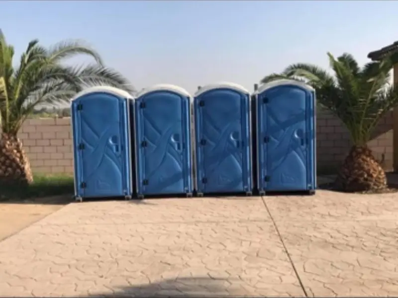 Four blue standard portable toilets lined up on a stamped-concrete patio between palm trees